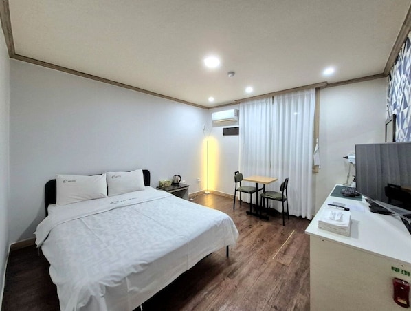 A hotel room with a bed, a desk, a chair, a small table, and an air conditioning unit.