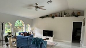 A spacious living room with a blue sofa, a ceiling fan, a flat-screen TV, and a large window with arched tops. - Charming 3-bedroom house with AC, WiFi in beautiful Naples (Naples)