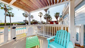 A covered porch with a pool, white and turquoise Adirondack chairs, and a view of palm trees and houses. - Charming Townhouse,  Shared pool, Great Location (Port Aransas)