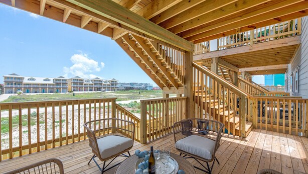 A wooden deck with wicker chairs and a table, overlooking a beach and buildings. - Private Heated Pool, Golf Cart Included, Short Walk to the Beach (Port Aransas)
