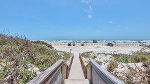 A beach with a wooden staircase leading down to the sand, a lifeguard tower, and vehicles parked along the beach. - Private Heated Pool, Golf Cart Included, Short Walk to the Beach (Port Aransas)