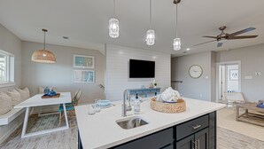 A modern kitchen with a central island, pendant lights, a ceiling fan, and a flat-screen TV on the wall. - Private Heated Pool, Golf Cart Included, Short Walk to the Beach (Port Aransas)