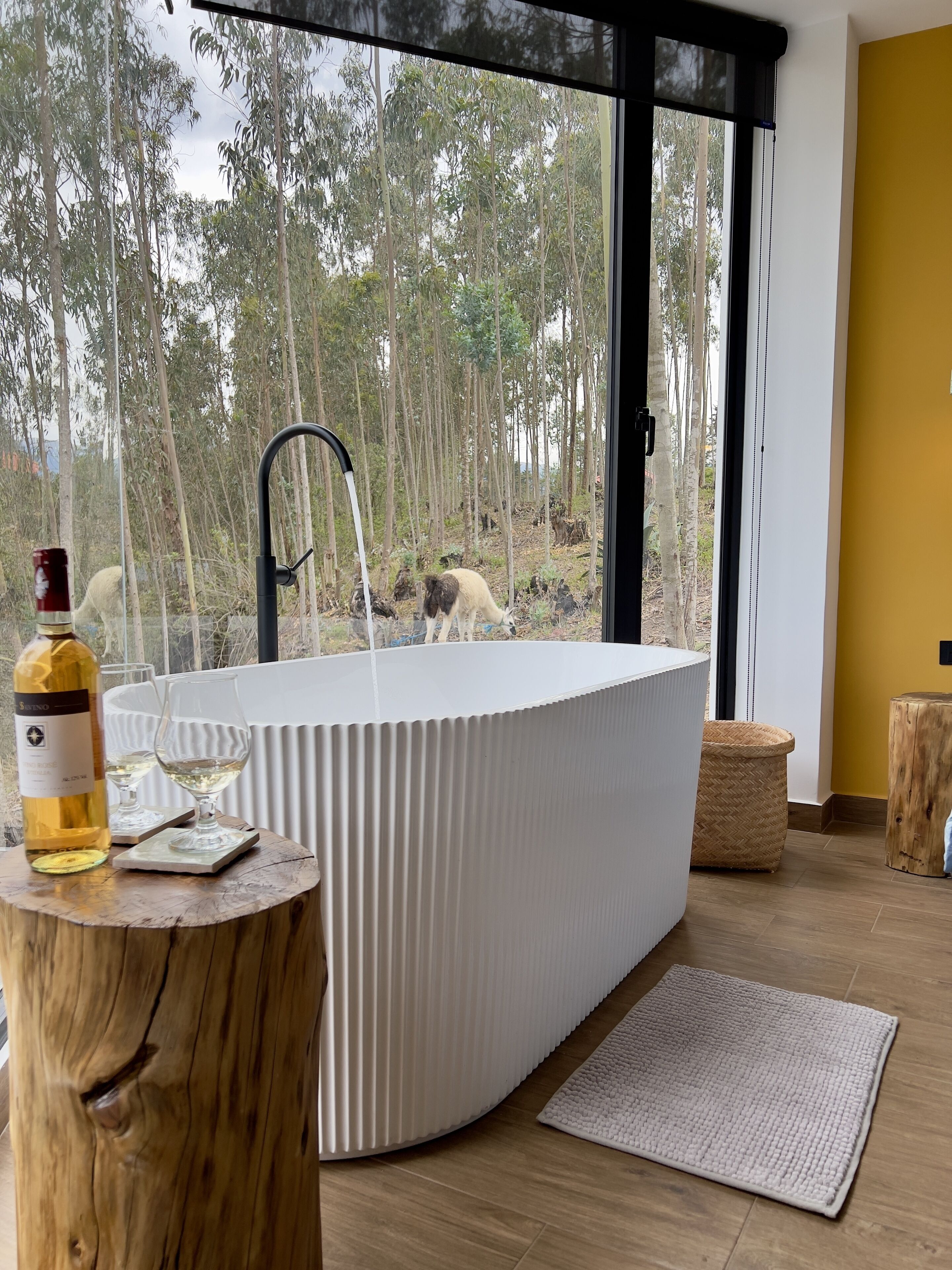 Un baño moderno con una bañera independiente, un taburete de madera y una botella de vino sobre un tocón de árbol.