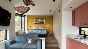Un interior moderno con un sofá, una mesita con un frutero, una cama con ropa de cama floral y una cocina con armarios rosas.