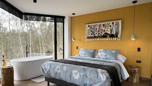 Un dormitorio moderno con una cama grande, una bañera y vistas a árboles a través de amplios ventanales.