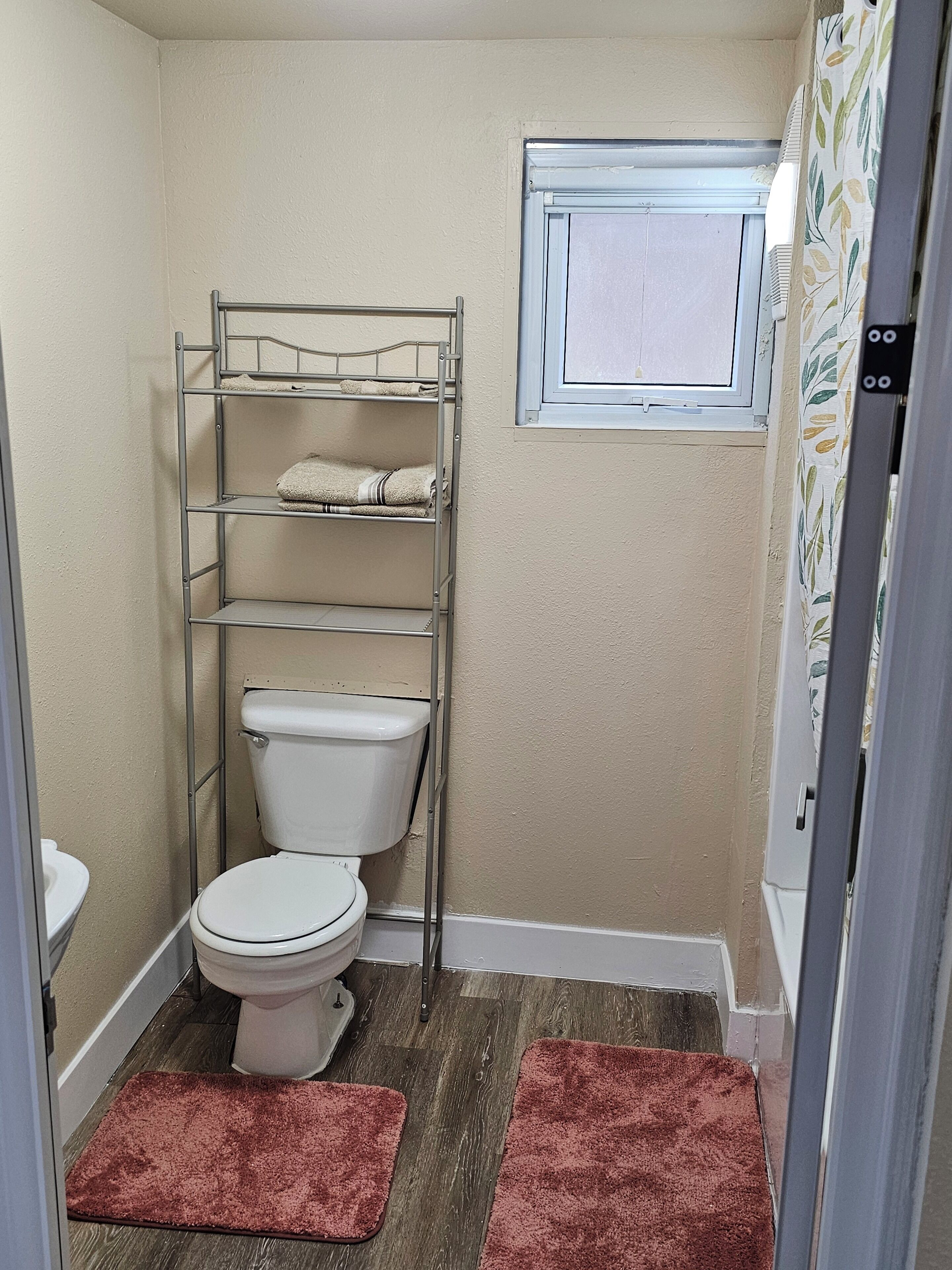 Ein kleines Badezimmer mit Toilette, einem Metallregal mit Handtüchern, einem Fenster und Holzboden.