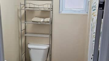 Ein kleines Badezimmer mit Toilette, einem Metallregal mit Handtüchern, einem Fenster und Holzboden.