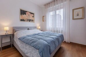 A bedroom with a bed, two bedside tables, a nightstand, a window with sheer curtains, and wall art.