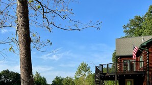 A hammock tied to a tree by a lake, a wooden cabin with an American flag, and a deck with outdoor furniture. - Secluded 5-Bedroom Lodge with Private Lake – Perfect for Large Groups! (Perryville)