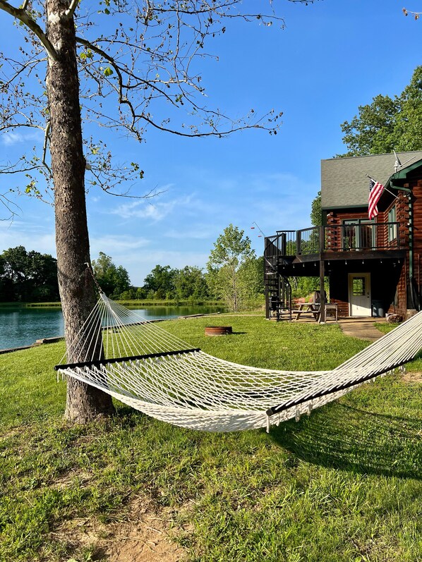 A hammock tied to a tree by a lake, a wooden cabin with an American flag, and a deck with outdoor furniture.