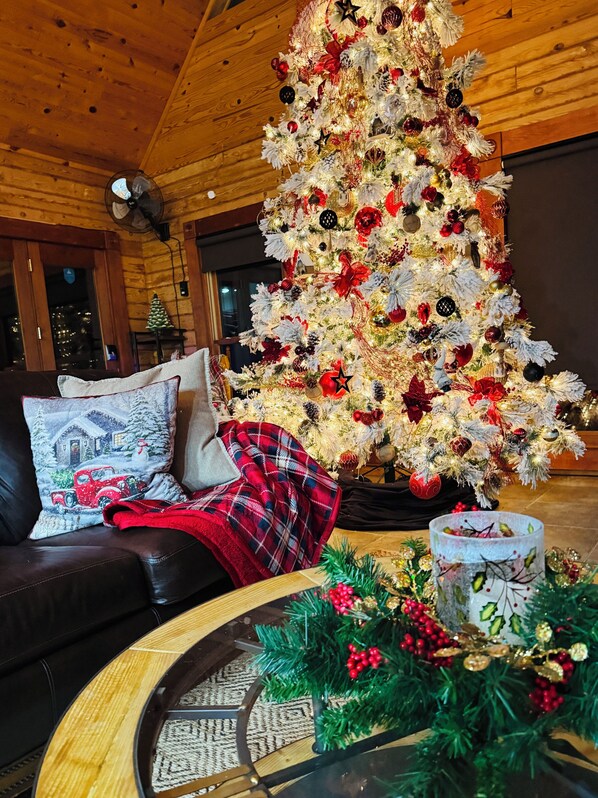 Ein gemütliches Wohnzimmer mit einem großen, geschmückten Weihnachtsbaum, einer Ledersofa mit karierter Decke und einem Couchtisch mit Glasdekoration.