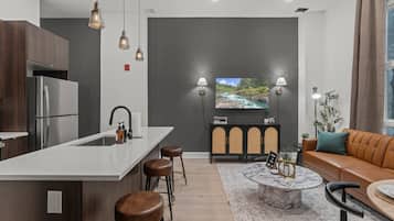 A modern kitchen with a central island, bar stools, a marble coffee table, and a sofa in a living area with a TV and plants.