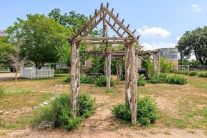 A wooden arbor structure in a garden setting with a dirt path leading to it, surrounded by greenery and a white fence. - Baron's Creek Ranch | Big Groups | 3 Homes (Fredericksburg)