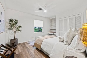 A bedroom with a bed, a bedside table, a chair, a potted plant, a window with a view, and a ceiling fan.