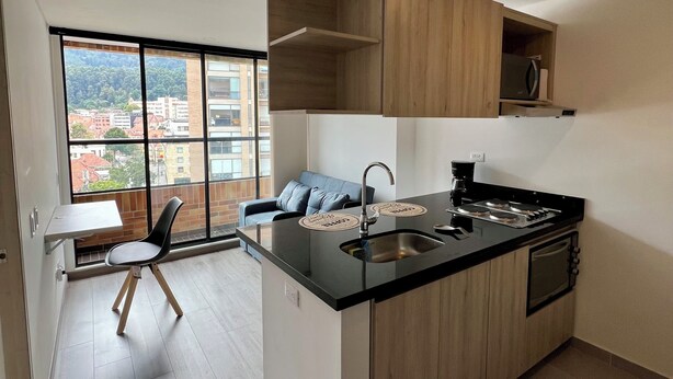 A modern kitchen with a black countertop, wooden cabinets, and a built-in stove. There is a window providing a view of buildings and trees. - Welcome to your perfect getaway in the heart of Bogota (Bogotá)