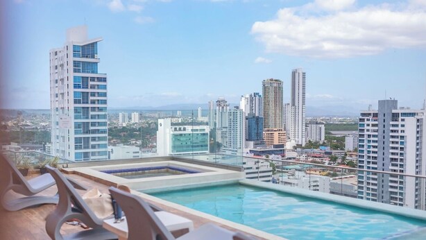 Une piscine à débordement située sur le toit, avec des chaises longues, offrant une vue panoramique sur la ville et ses gratte-ciel.