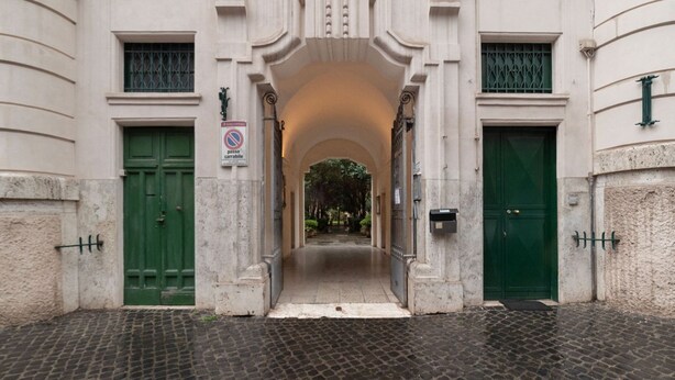 Entrada de um edifício com duas portas verdes, placa de proibição de estacionamento e um caminho de pedras que leva a uma área coberta.