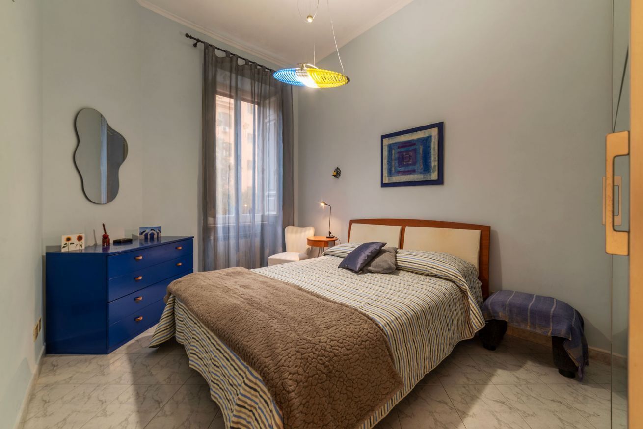 Une chambre à coucher avec un lit, une commode bleue, une table de chevet, une lampe, une fenêtre avec des rideaux et un miroir fixé au mur.