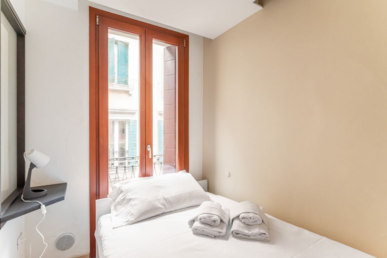 Une chambre d’hôtel avec un lit, un bureau doté d’une lampe, une fenêtre donnant sur des immeubles et une paire de serviettes sur le lit.
