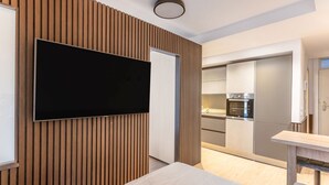 Ein modernes Hotelzimmer mit einem an der Wand befestigten Flachbildfernseher, einer Wandverkleidung aus Holzpaneelen, einem Bett, einem kleinen Tisch und einer Küchenzeile im Hintergrund.