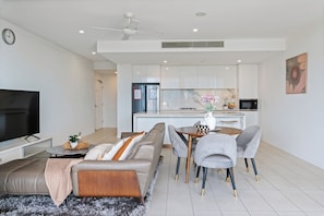 A modern living room with a sofa, a coffee table, and a TV. There is a dining area with a table and chairs. The kitchen is visible in the background with white cabinets and a microwave.