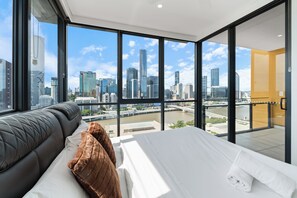 A modern hotel room with a large window offering a city view, a bed with white linens, and a grey sofa.