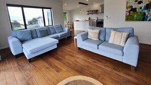 A modern living room with two light blue sofas, wooden flooring, a large window, and a kitchen in the background. - Riverton Luxury in the Lane (Riverton / Aparima)
