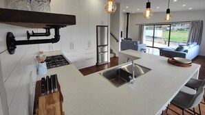 A modern kitchen with white cabinets, a stainless steel refrigerator, and a central island with a sink. There are hanging pendant lights and a view of the outdoors through large windows. - Riverton Luxury in the Lane (Riverton / Aparima)