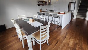 A modern kitchen with white cabinetry, a central island, and a dining area with a dark wooden table and white chairs. - Riverton Luxury in the Lane (Riverton / Aparima)