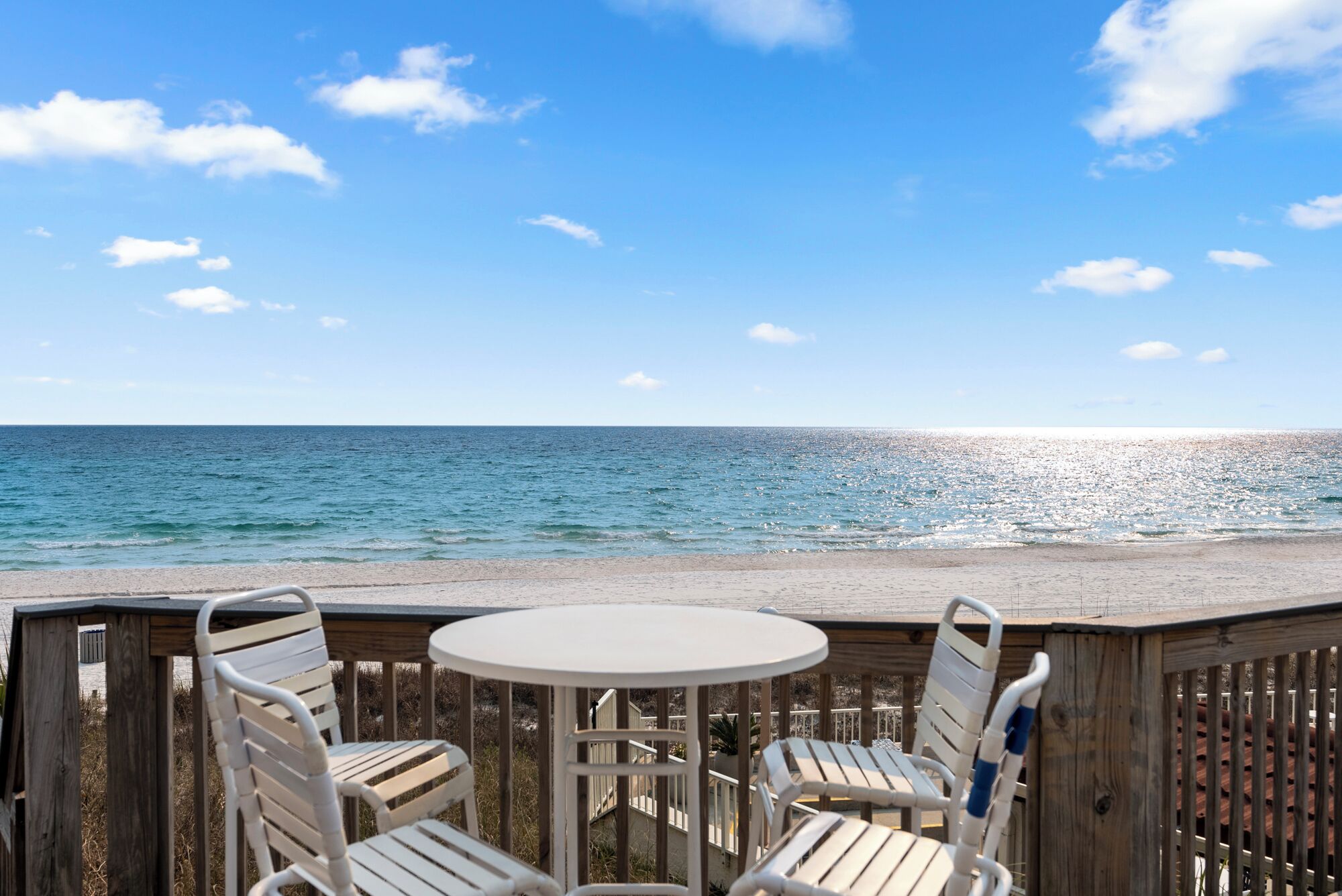 Terraza de madera con una mesa redonda y dos sillas, con vistas a una playa, el océano y un cielo despejado.