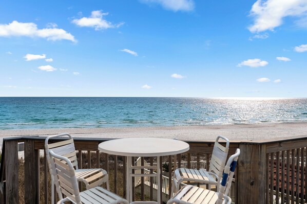 A wooden deck with a round table and two chairs, overlooking a beach with the ocean and a clear sky.