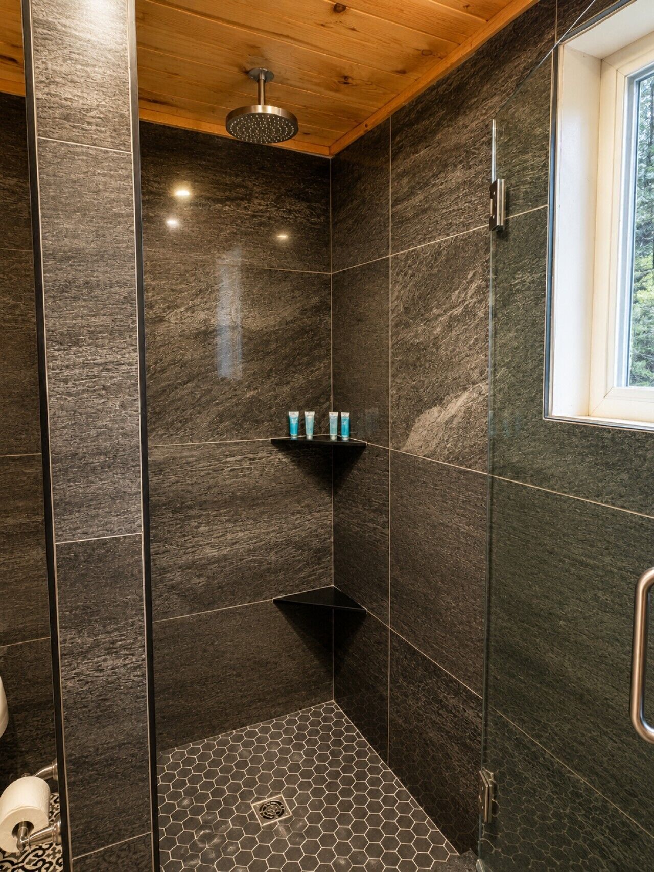 Un espace douche moderne avec des carreaux de couleur sombre, une pomme de douche et une étagère contenant des articles de toilette.