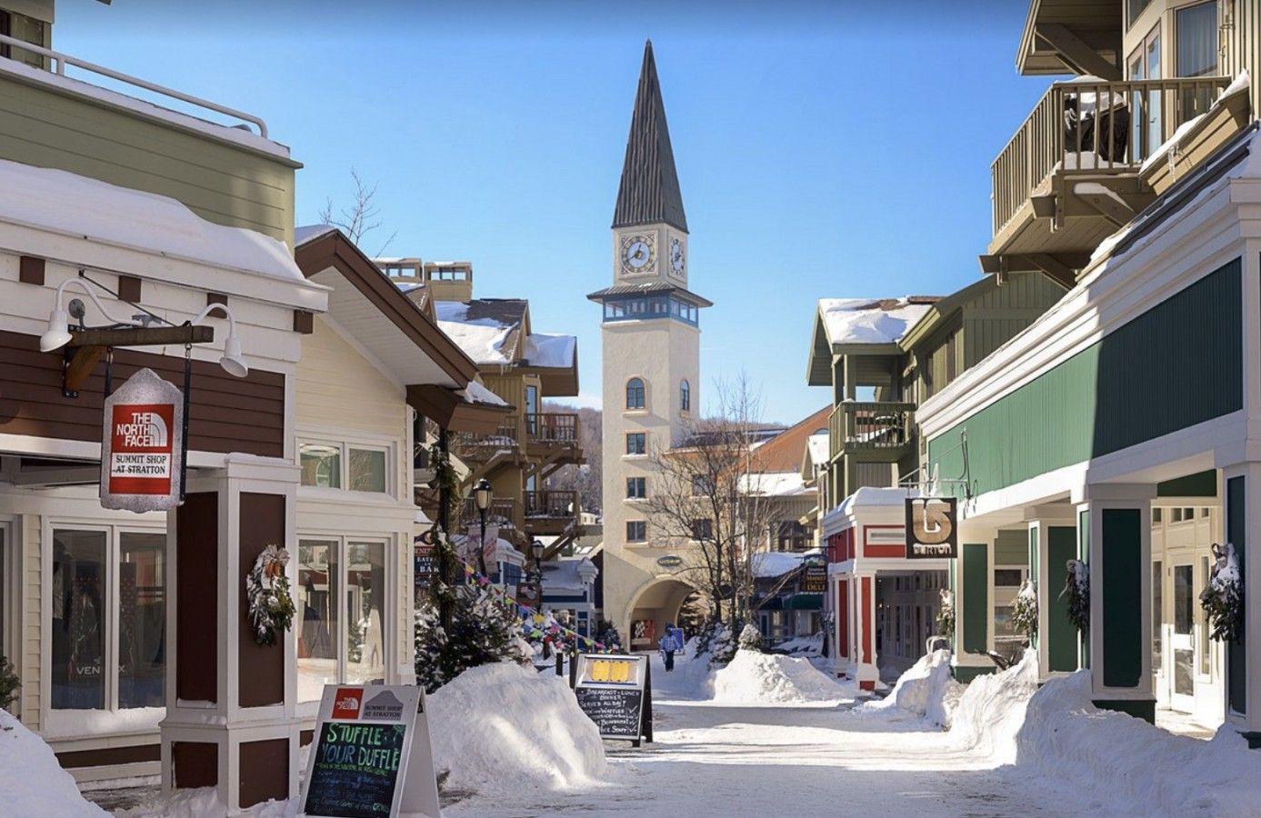 Une rue enneigée bordée d’immeubles, avec un beffroi et une enseigne indiquant « The North Face Summit Shop ».