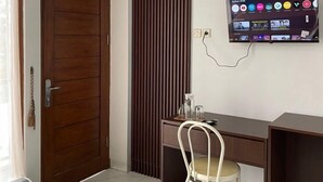 A modern room with a wooden door, a tall vertical panel, a desk with a chair, and a mounted TV displaying various channels. - Laika Homestay (Banguntapan)