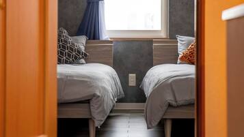 Un couloir étroit mène à une chambre dotée de deux lits, d’une fenêtre avec des rideaux et d’un plancher en bois.