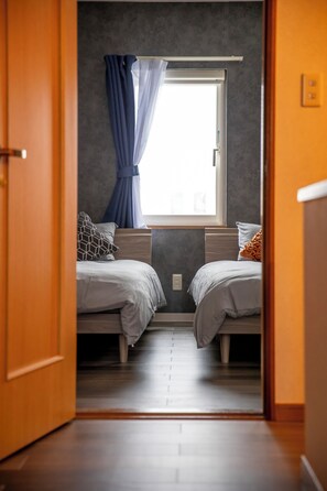 A narrow hallway leading to a bedroom with two beds, a window with curtains, and a wooden floor.