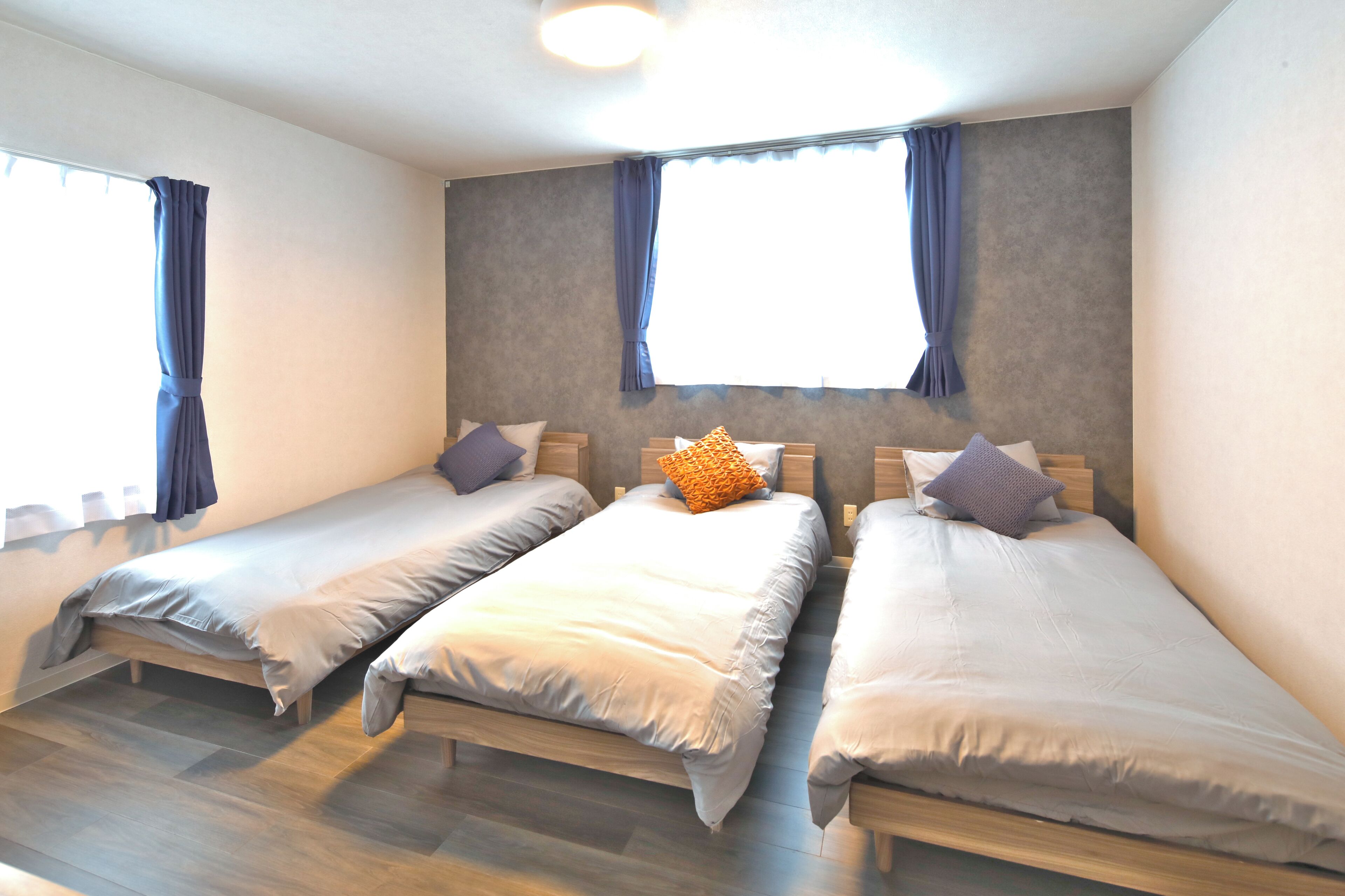 A room with three single beds, each with a pillow and a blanket. There are two windows with curtains, and the room has a wooden floor.