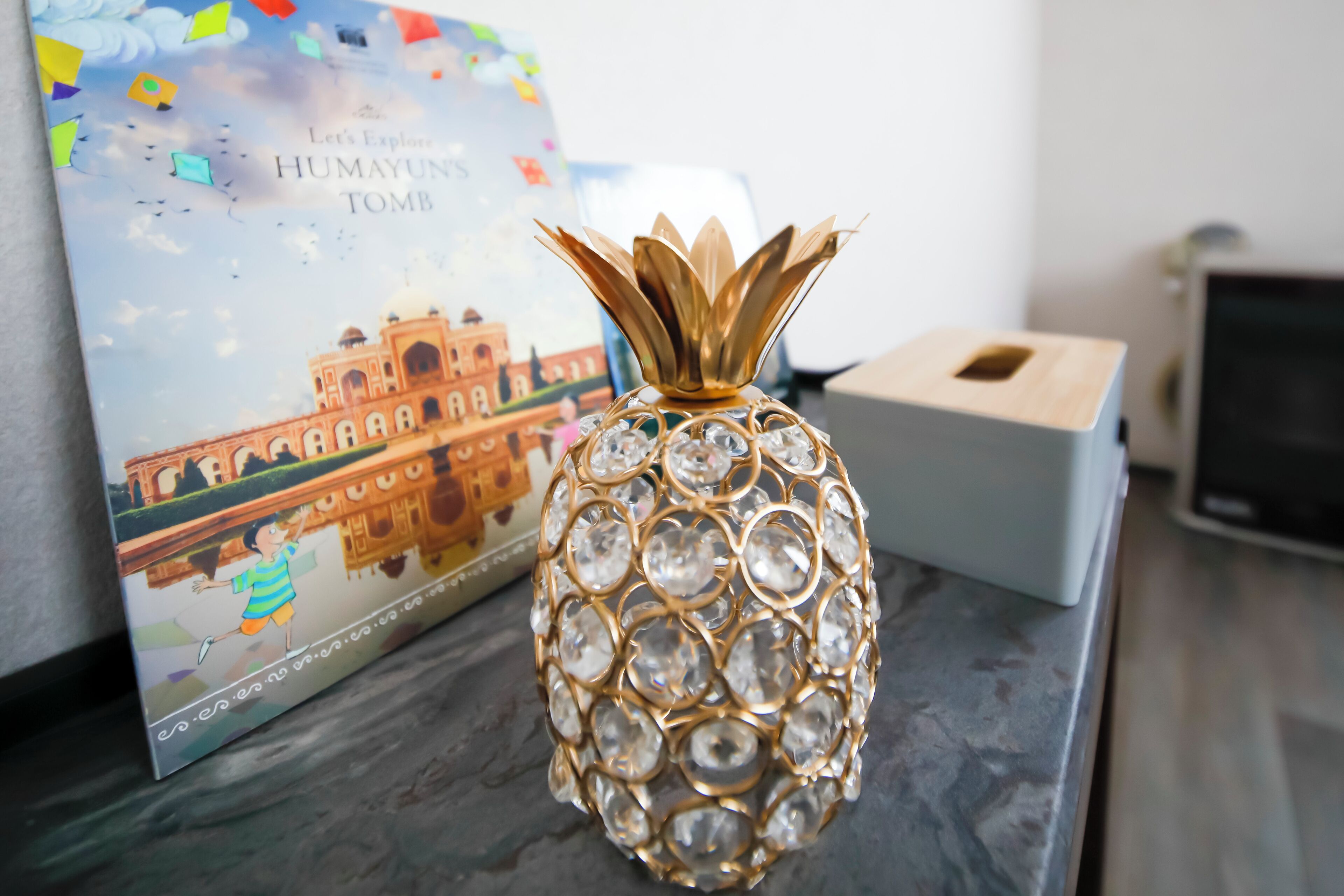 A decorative pineapple-shaped vase with a gold and crystal design, placed on a table next to a book titled "Let's Explore Humayun's Tomb".