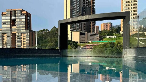 A modern building with a glass facade, a reflection pool, and surrounding high-rise buildings. - 1002 - Beautiful Apartment In El Poblado Medellin (Medellín)