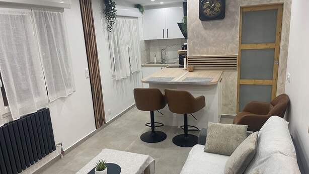 Un moderno salón con un sofá, dos sillones, una mesa de centro con una planta, una zona de cocina con encimera y armarios, y un reloj en la pared.