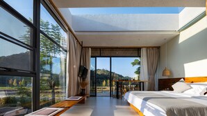 A modern bedroom with a large bed, a desk, and a view of a mountainous landscape through large windows. - Yingde YeDuGu Resort (Qingyuan)