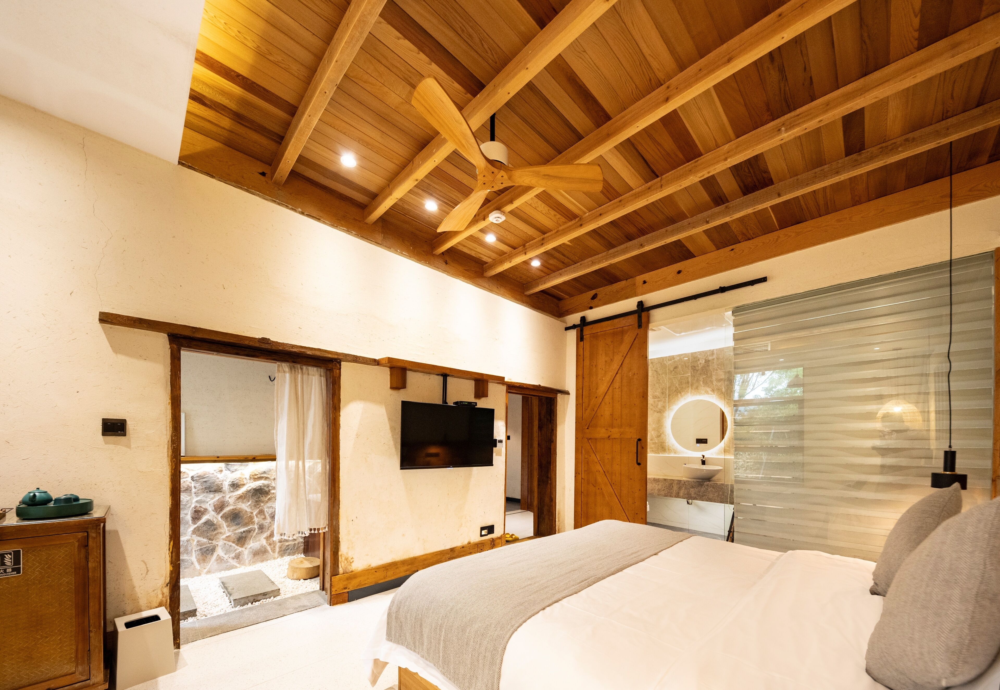 Un dormitorio con techo de madera, una cama, un televisor, un baño con espejo y una pared de piedra.