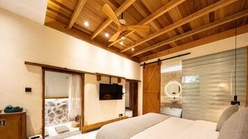 Un dormitorio con techo de madera, una cama, un televisor, un baño con espejo y una pared de piedra.