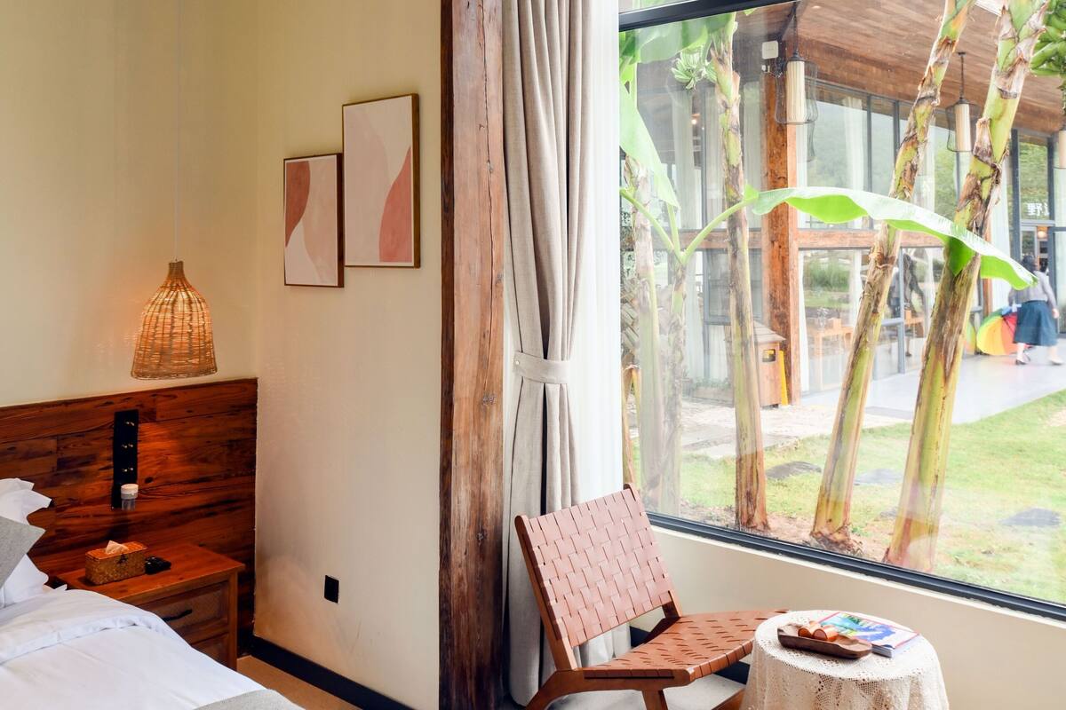 Ein Schlafzimmer mit einem Bett, einem Nachttisch aus Holz, einem Sessel, einem kleinen Tisch und einem großen Fenster mit Blick ins Freie.
