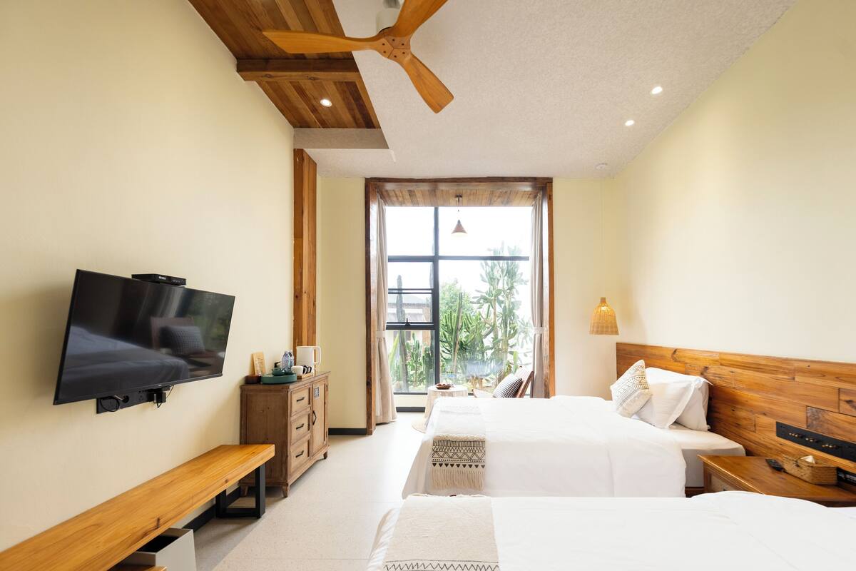Ein Hotelzimmer mit zwei Betten, einem Fernseher, Holzmöbeln und einem großen Fenster mit Blick auf Grünflächen.
