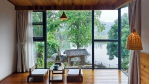 A room with wooden flooring, a table with a teapot, two chairs, and a large window offering a view of a traditional house and greenery. - Yingde YeDuGu Resort (Qingyuan)