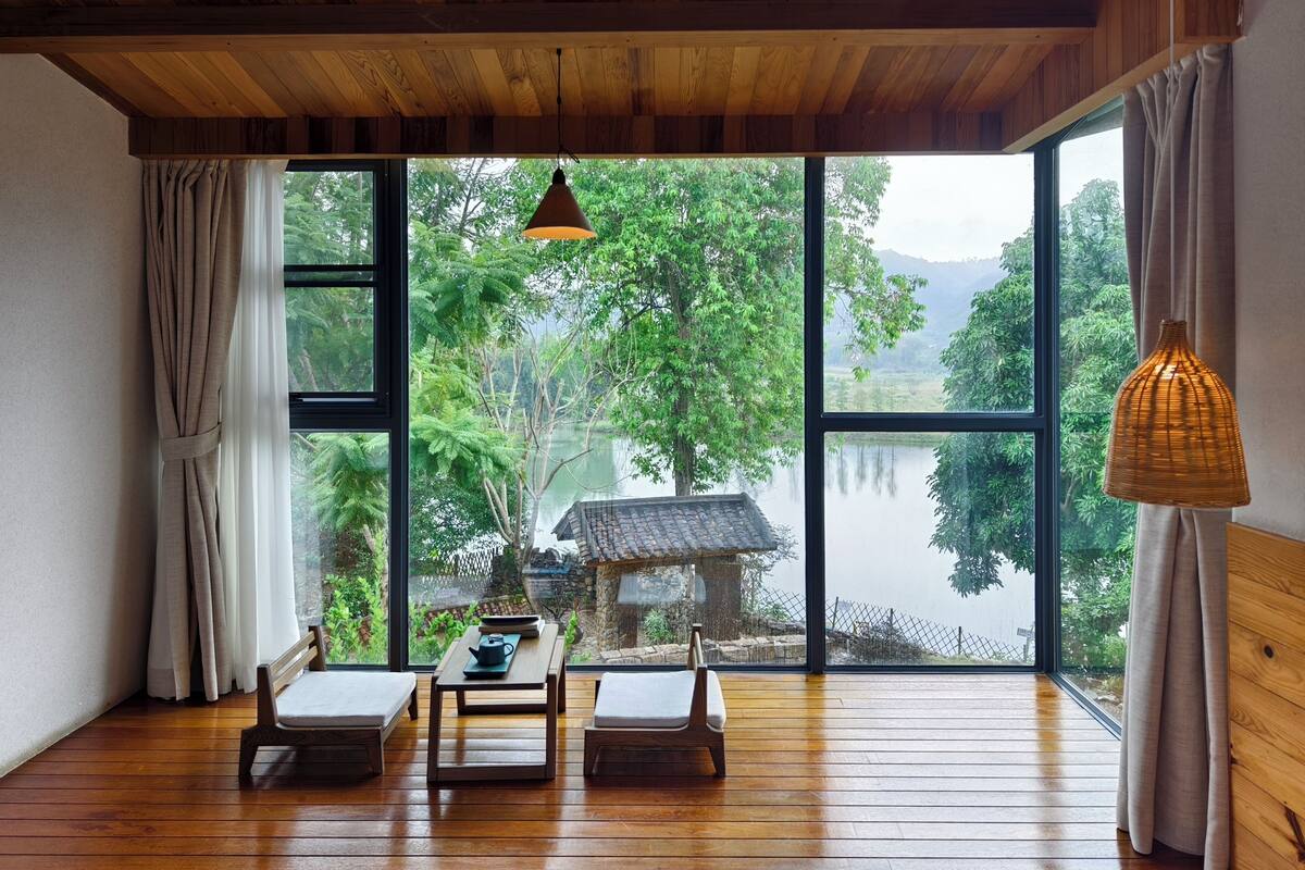 Ein Zimmer mit Holzboden, einem Tisch mit Teekanne, zwei Stühlen und einem großen Fenster mit Blick auf ein traditionelles Haus und Grünflächen.