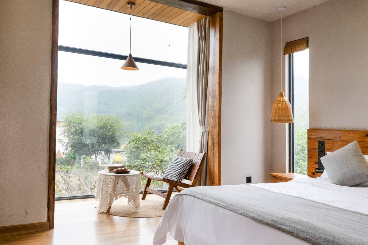 Ein Schlafzimmer mit großem Fenster, einem Bett, einem Sessel und einem kleinen Tisch mit Obstschale.