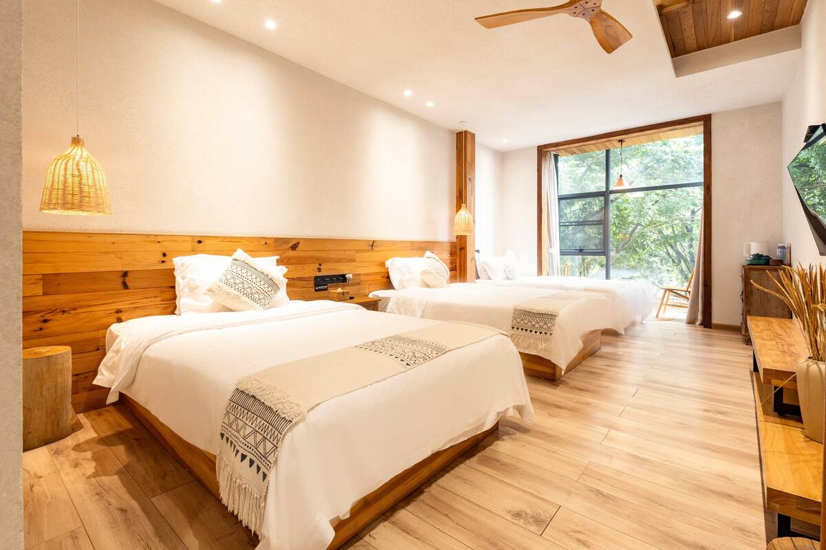 Ein Hotelzimmer mit zwei Betten, Holz-Kopfteilen, einem Deckenventilator und einem großen Fenster mit Blick auf Bäume.