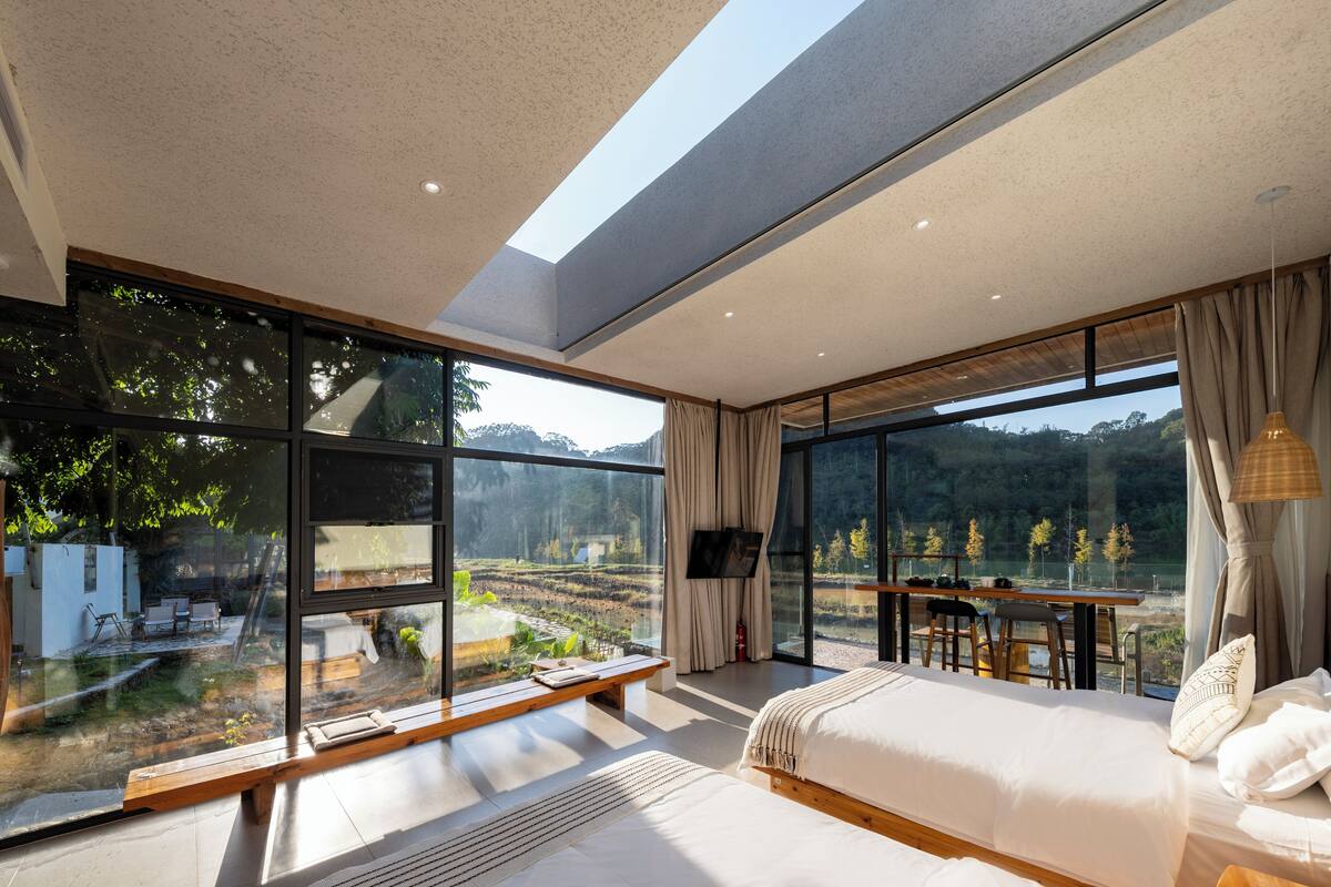 Ein modernes Schlafzimmer mit großen Fenstern, einem Bett, einem Schreibtisch und Blick auf eine bewaldete Landschaft.